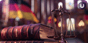 A set of scales and a stack of books sit on a courtroom desk with a German flag waving in the background