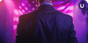 A white man in a suit with his back to the camera faces an American flag