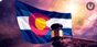 A Colorado flag waves in front of a sunset; a gavel on a sounding block rests in front of the flag