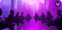 The silhouettes of a group of 9 figures sit at a table in a conference room with floor to ceiling windows overlooking a skyline of skyscrapers at sunset